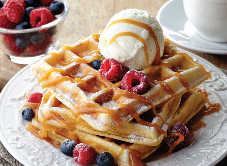 Waffles topped with caramel sauce and ice cream. fresh berries scattered across the dessert and in a pot to the side